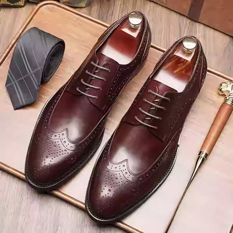 Pair of brown leather dress shoes on a wooden surface with a tie and pen.