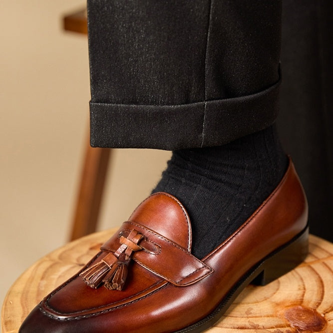 Brown leather loafer with tassels worn with black pants and socks on a wooden stool.