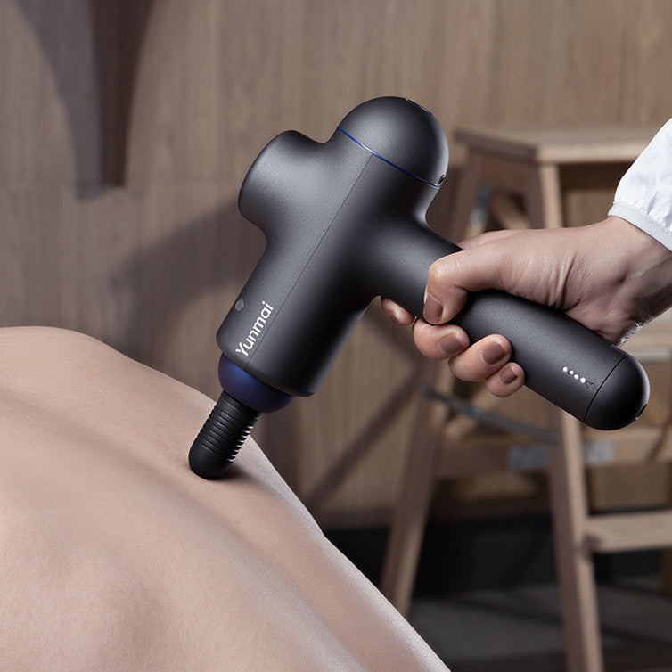 Person using a black massage gun on their arm with a blurred indoor background