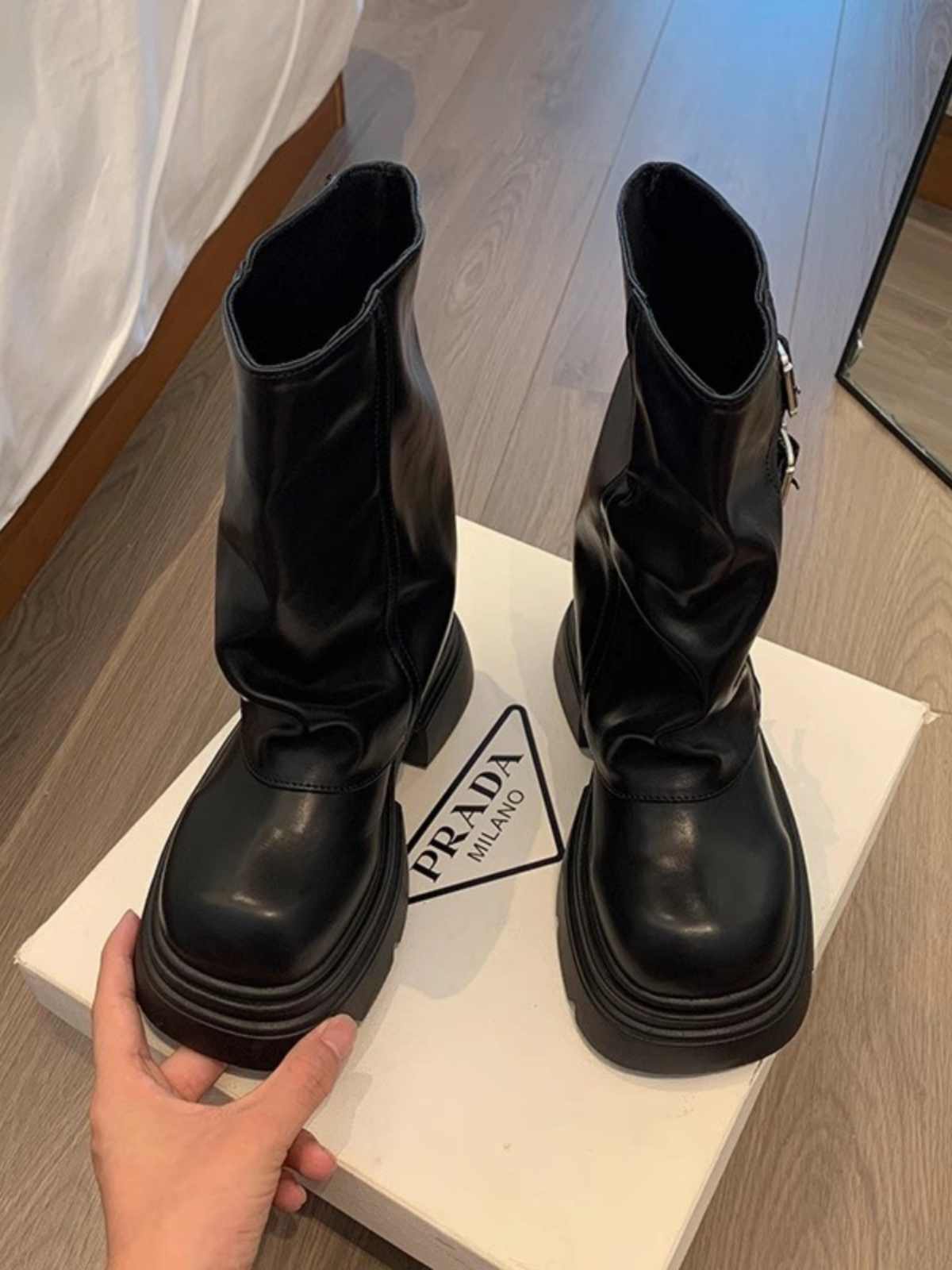 Black boots on a Prada box with a hand holding one of the boots.
