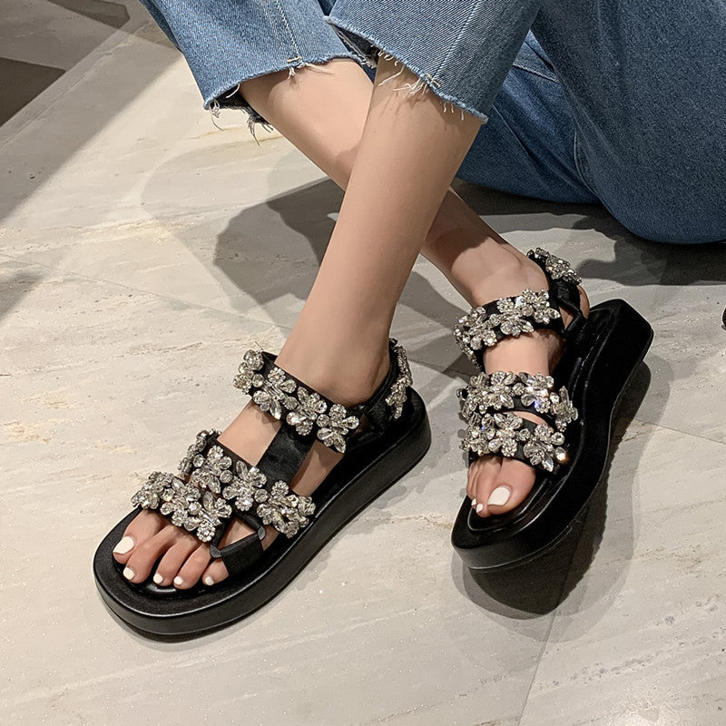Black sandals with silver embellishments worn by a person on a light wooden floor.