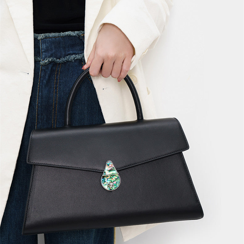 Black handbag with a decorative clasp held by a person wearing a white coat and blue jeans.