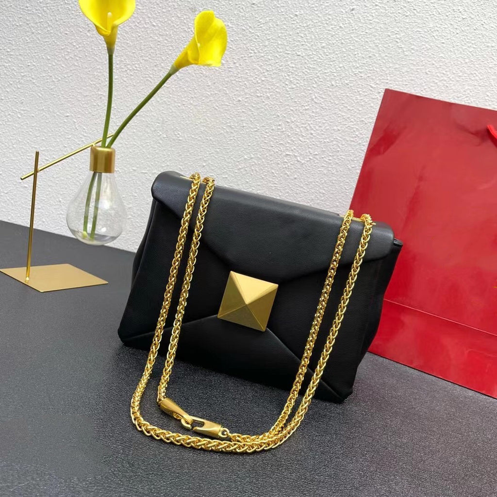 Black handbag with gold chain and pyramid detail on a gray surface with yellow flowers and a red bag in the background.