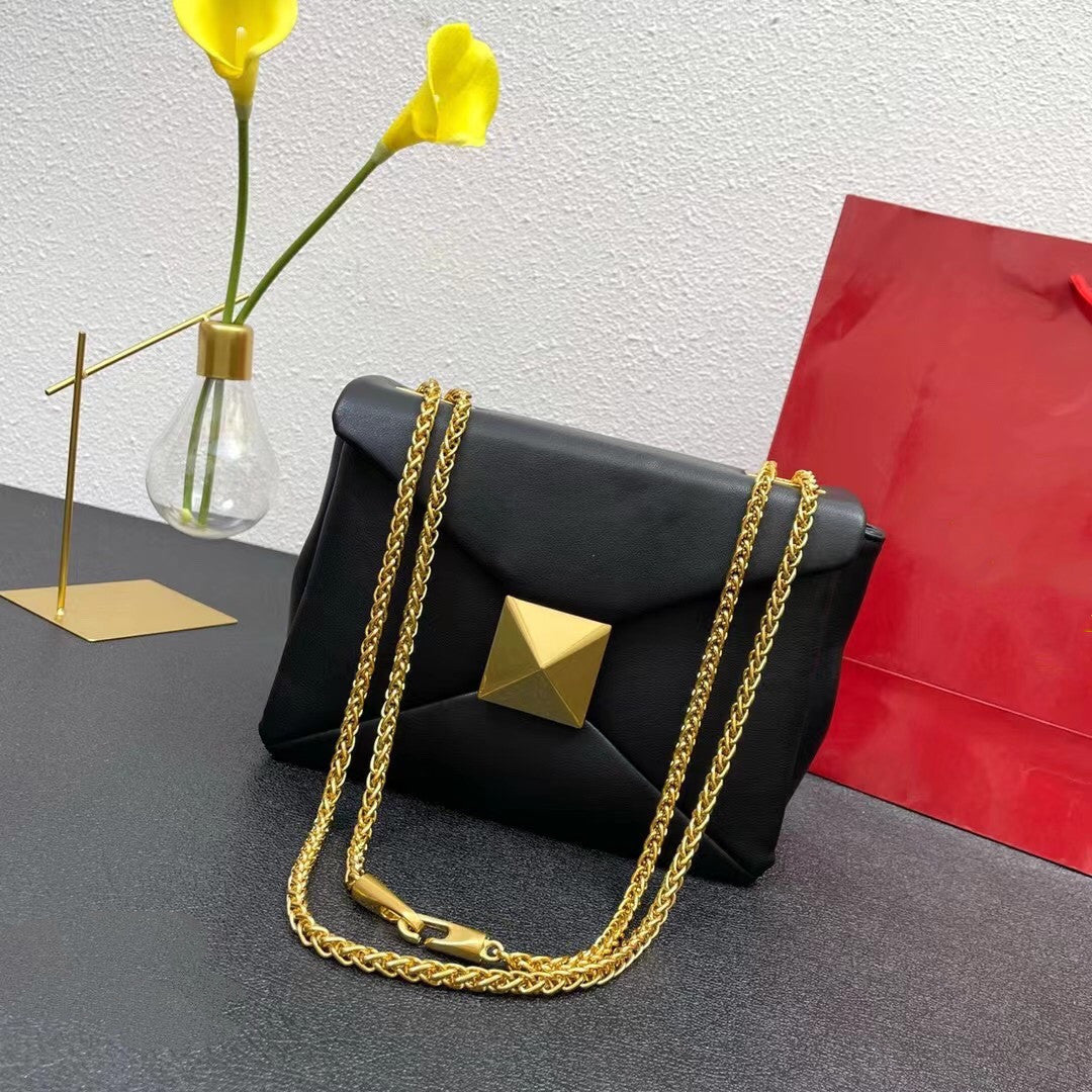 Black handbag with gold chain and pyramid detail on a gray surface with yellow flowers and a red bag in the background.