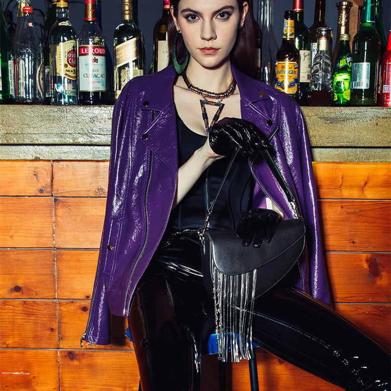 Woman in a purple coat holding a black handbag in front of a bar with bottles on shelves.