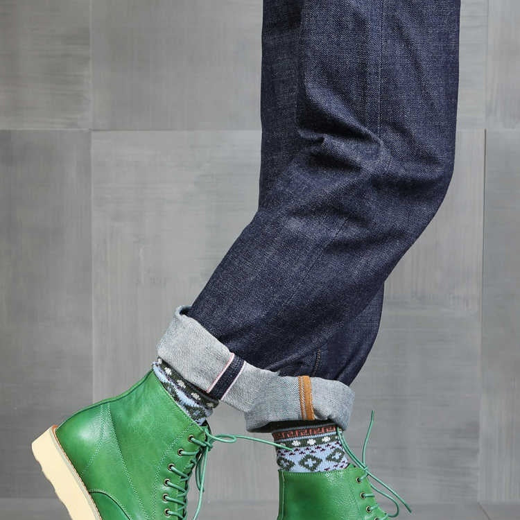 Green leather boots with beige soles worn with blue jeans on a gray wooden floor.