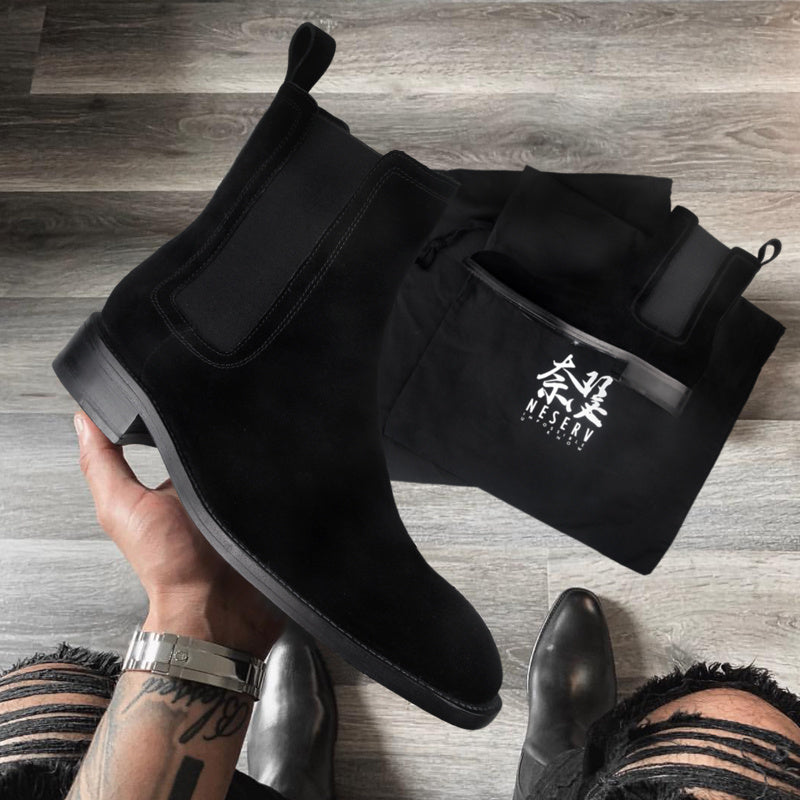 Black Chelsea boots held by a hand on a wooden floor with a black shoe bag featuring a logo.