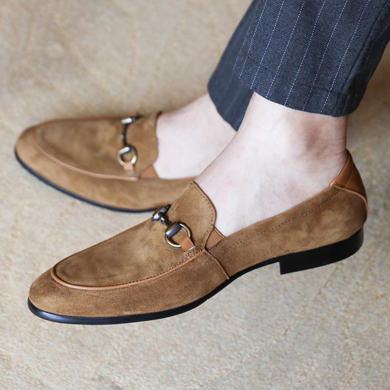 Brown suede loafers with a metallic buckle worn by a person on a neutral background
