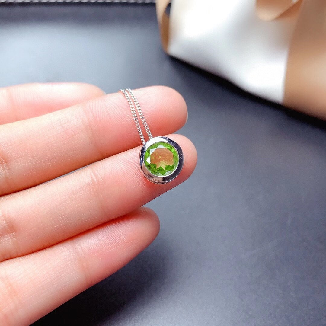 Silver necklace with a green gemstone pendant held between fingers on a dark surface.