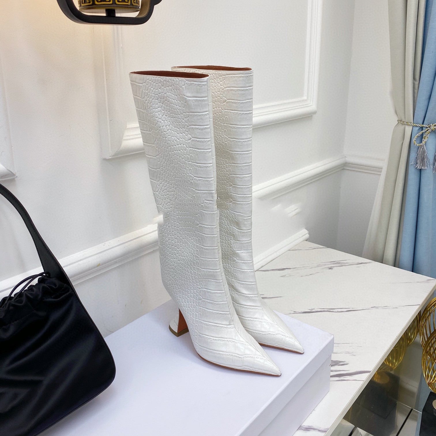 White textured knee-high boots on a white platform with a black handbag beside them.