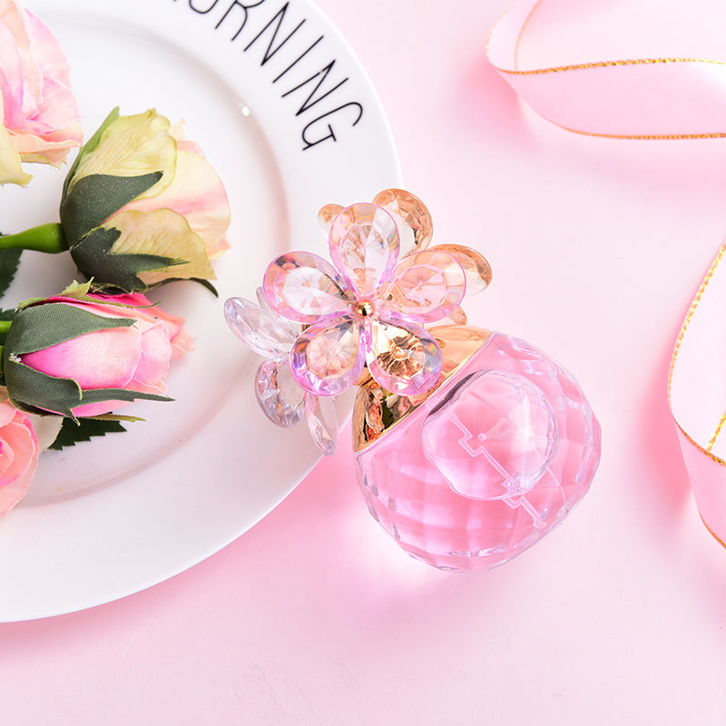 Pink perfume bottle with floral design on a pink surface with flowers and a plate in the background.