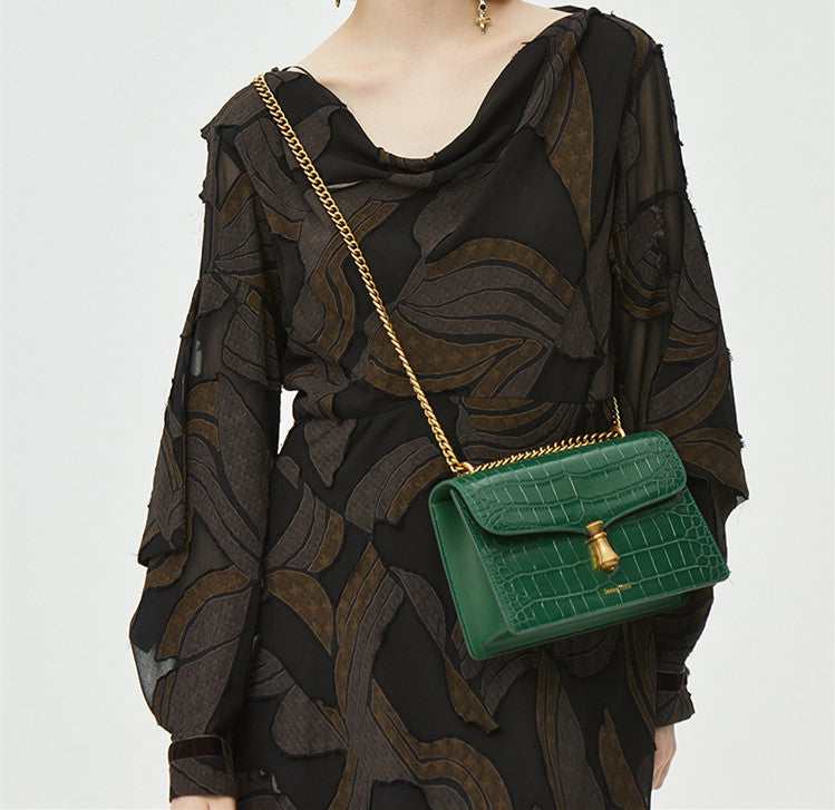Green handbag with gold chain strap worn by a person in a black patterned outfit.