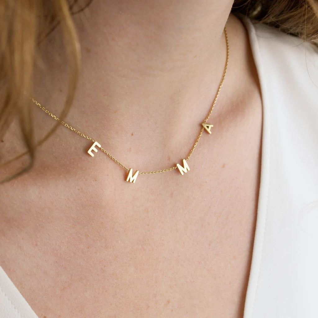 Gold necklace with 'EMMA' letters on a person wearing a white top.
