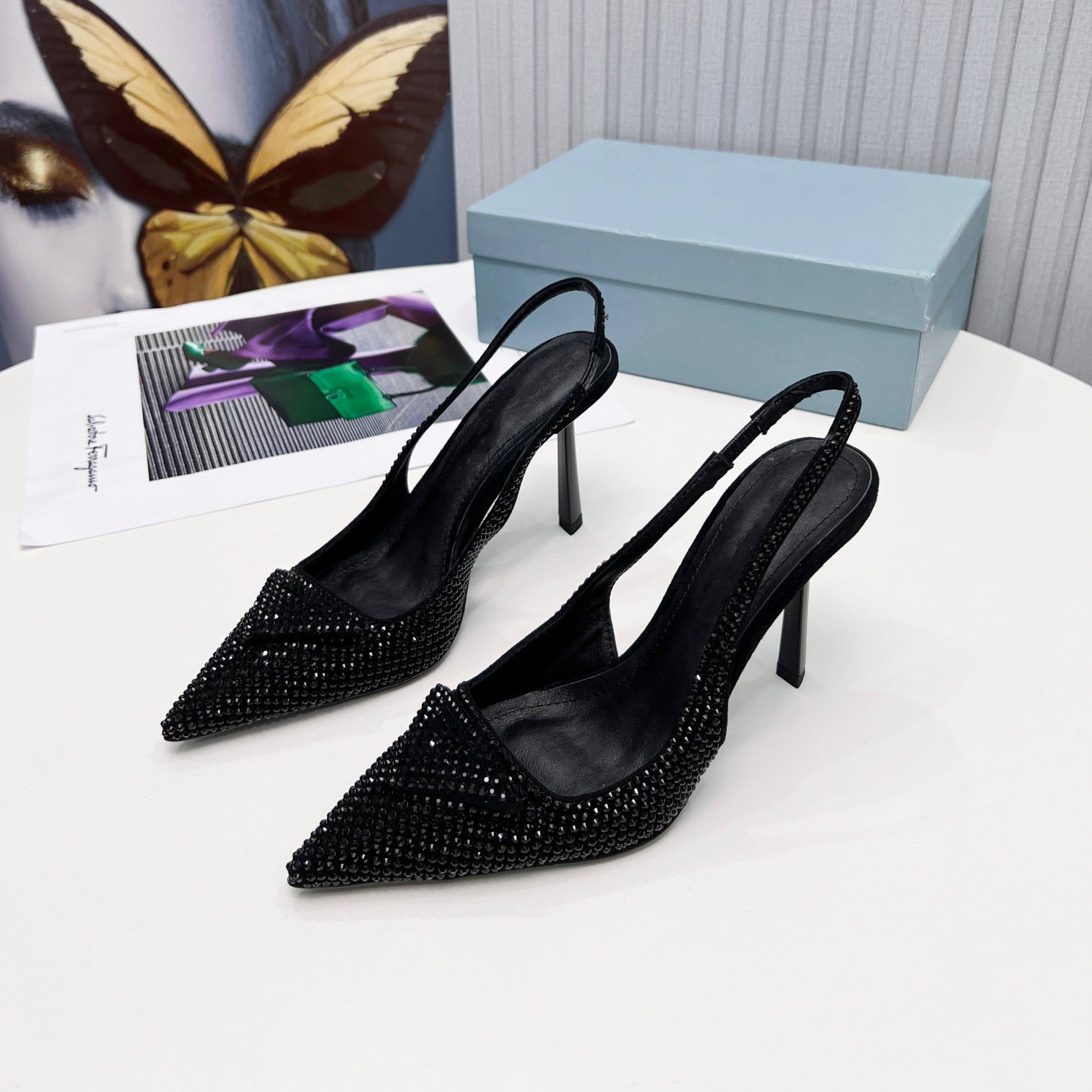 Black stiletto shoes with crystal embellishments on a white surface with a butterfly and magazine in the background.