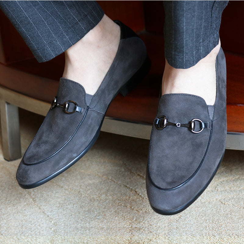 Gray suede loafers with a metallic buckle worn by a person sitting on a chair.