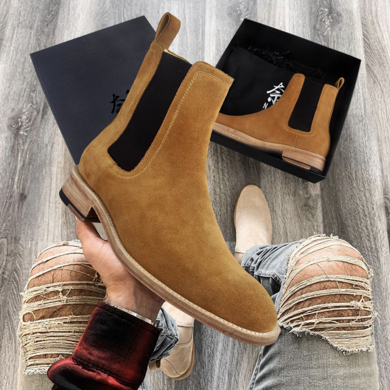 Brown suede Chelsea boots held by a person on a wooden floor with black boxes in the background.