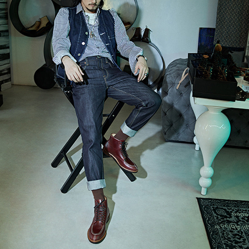 Person sitting on a chair wearing a checkered shirt, vest, jeans, and brown shoes in an indoor setting.