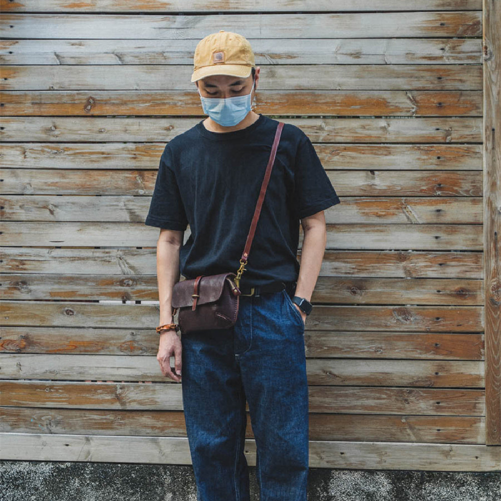 Person wearing a black t-shirt, blue jeans, and a yellow cap standing against a wooden wall.