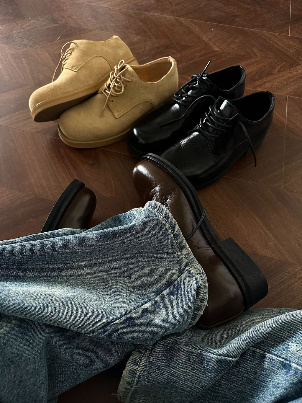 Pair of beige shoes, black shoes, and brown shoes on a wooden floor with jeans.