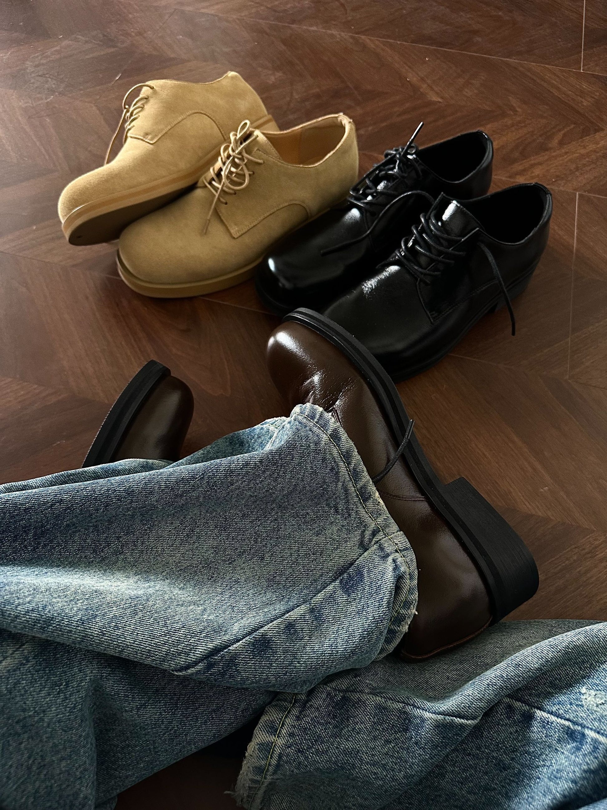 Pair of beige shoes, black shoes, and brown shoes on a wooden floor with jeans.