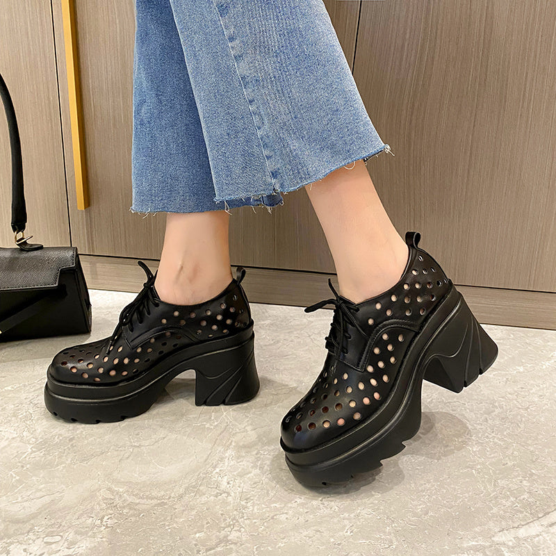 Black perforated platform shoes worn with blue jeans on a light gray floor.