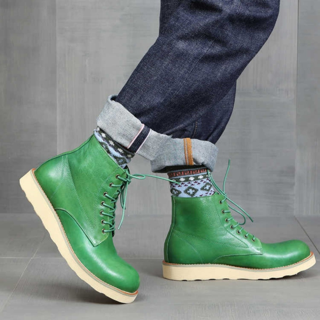 Green leather boots worn with blue jeans and patterned socks on a gray floor.