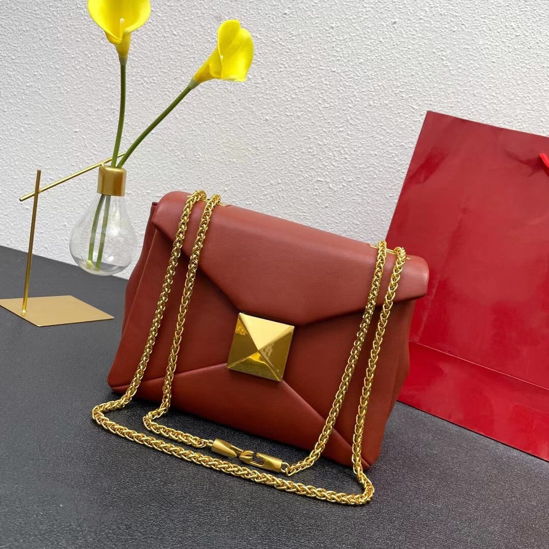 Red handbag with gold chain and clasp on a gray surface with yellow flowers and a red bag in the background.
