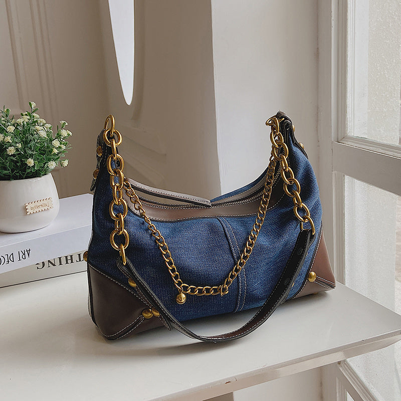 Blue handbag with gold chain on a white surface, with a plant and books in the background.